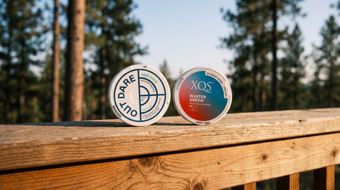 Two nicotine pouch cans resting side by side on a weathered cedar deck railing outdoors