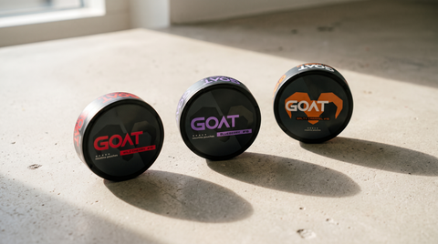 Three GOAT nicotine pouch cans in different flavours lined up on a pale concrete surface