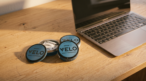 Round nicotine pouch cans arranged beside an open laptop on a warm oak desk