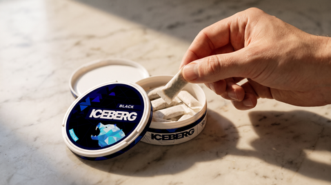 A hand lifting a nicotine pouch from an open round flat pouch can resting on a marble worktop