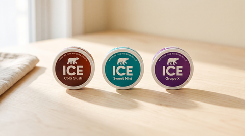 ICE nicotine pouch cans lined up on pale birch wood, Cola Slush, Sweet Mint and Grape X facing forward