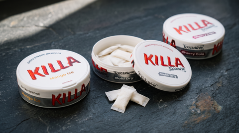 Three KILLA nicotine pouch cans lined up on dark slate, centre can open showing pouches