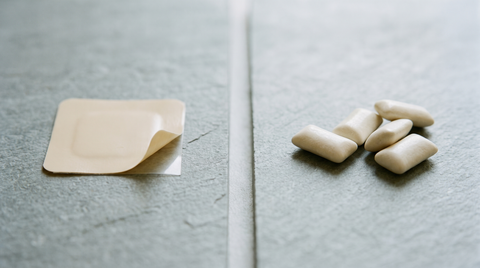 An unused nicotine patch and unwrapped gum pieces side by side on a pale slate surface