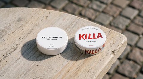 Two nicotine pouch cans side by side on a sandstone café table with cobblestones behind
