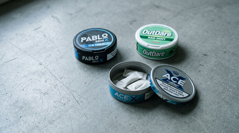 Three nicotine pouch cans of different brands clustered on a worktop, one open showing white pouches