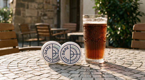 Two nicotine pouch cans on a cobblestone café table beside a glass of dark lager