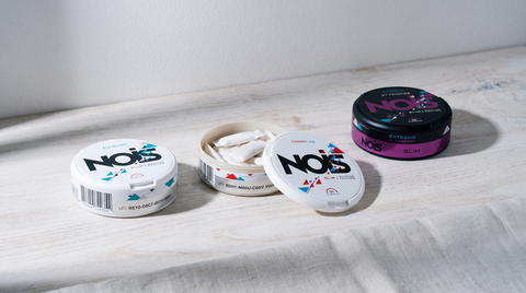 Three NOIS nicotine pouch cans arranged on a pale birch surface, one open showing white pouches