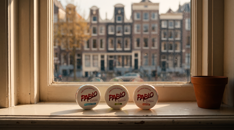 Three Pablo Exclusive nicotine pouch cans on a Dutch apartment windowsill with canal houses softly blurred outside