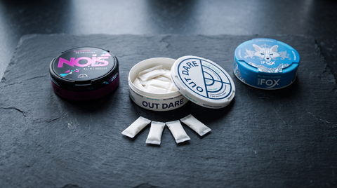 Three nicotine pouch cans in a row on slate, centre can open showing white pouches inside