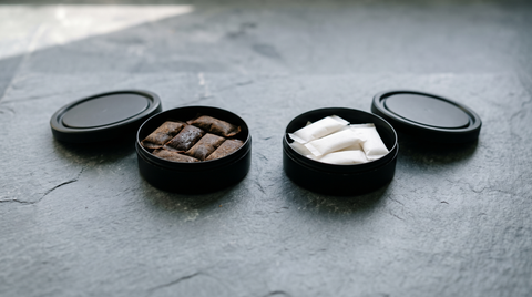 Two open plain cans side by side, one holding dark snus portions and one holding white nicotine pouches