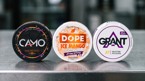 Three round nicotine pouches with CAMO, DOPE and GRANT branding displayed on a grey metallic surface against a blurred backdrop