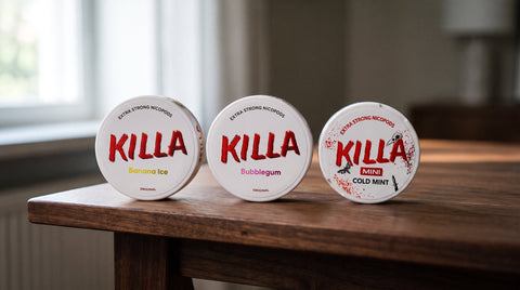 Three white KILLA nicotine pouch cans in Banana Ice, Bubblegum and Cold Mint on a wooden table
