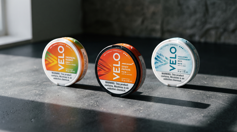Three VELO nicotine pouch cans in contrasting warm and cool colourways on dark concrete