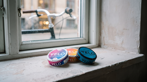 Three VELO nicotine pouch cans on a pale Dutch windowsill with a bicycle blurred softly outside