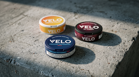 Three VELO nicotine pouch cans — Peppermint Max, Icy Cherry and Tropical Mango — on weathered concrete