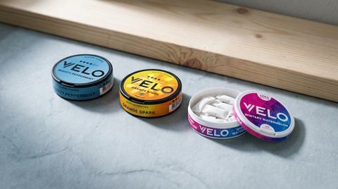 Three VELO nicotine pouch cans on pale grey slate, one open showing small white pouches inside