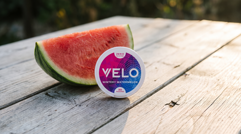 VELO Wintery Watermelon Zero nicotine pouch can beside a fresh watermelon slice on a wooden table