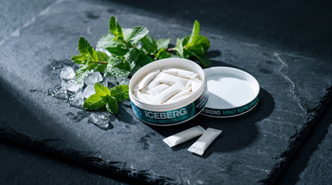Open nicotine pouch can with white pouches inside surrounded by mint sprigs and ice chips on dark slate