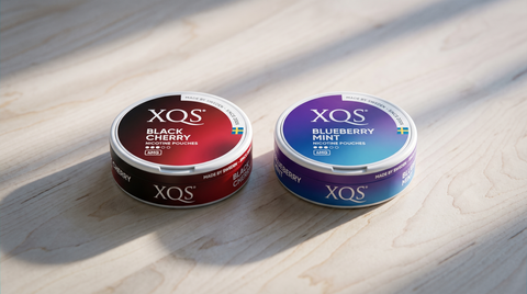 Two closed XQS nicotine pouch cans side by side on a pale birch-wood tabletop in cool Nordic light