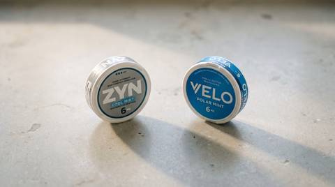 ZYN and VELO nicotine pouch cans placed side by side on a pale grey stone surface
