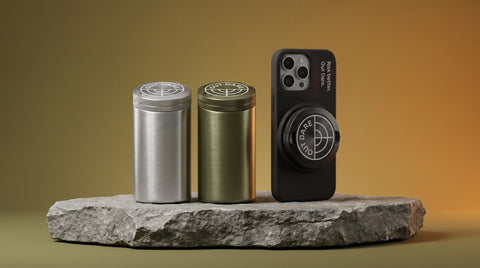 OutDare accessories line — brushed-metal silver and olive pouch cans beside a black iPhone case with the OUT DARE metal can mounted, on a stone slab
